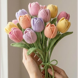 May include: A bouquet of handmade crochet tulips in various pastel colors, including pink, purple, yellow, and orange. The flowers have green stems and leaves, tied together with a green yarn bow. A craft item.