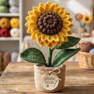 Puede incluir: Un girasol de ganchillo hecho a mano en una maceta tejida. El girasol presenta pétalos amarillos, un centro marrón, hojas verdes y un tallo verde. La maceta tiene una etiqueta que dice "Sunshine Stitches".