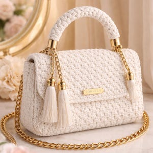 May include: Cream-colored crochet handbag with a gold chain strap and handle. The bag features gold accents, including the handle attachments and chain tassels. The word "LOVELLA" is displayed on a small gold plate on the front of the bag. The bag has a flap closure and a textured appearance.