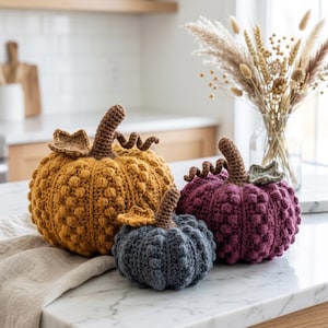 May include: Three handmade crochet pumpkins in mustard yellow, deep plum, and slate gray. Each pumpkin features a textured bobble stitch pattern, a brown crocheted stem, a curly tendril, and a small crocheted leaf attached to the top.