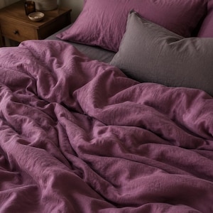 May include: A deep plum-colored duvet cover with a textured fabric finish, accompanied by matching plum pillowcases and a single dark gray rectangular pillow, all arranged on a bed with a soft, rumpled appearance.