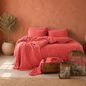 May include: A coral-coloured bed with matching bedding, including pillows and a duvet, dominates the scene. A wooden crate holds additional fabric. A potted plant and a decorative lantern with candles add to the room's ambiance. A rug is on the floor.