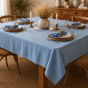 May include: A square light blue tablecloth draped over a wooden dining table. The table is set with white plates, blue cloth napkins in woven rings, clear glasses, silver forks, and two lit white candles in wooden holders with a central vase.