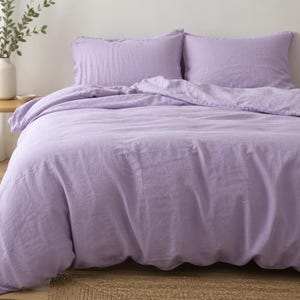 Puede incluir: Un juego de cama de color lavanda, que incluye una funda nórdica y dos fundas de almohada, se muestra en una cama. La ropa de cama tiene una textura suave y arrugada. Una pequeña mesa auxiliar de madera con un jarrón de vegetación está en el fondo.