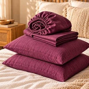 May include: A set of deep plum-colored bedding including two rectangular pillowcases and a folded flat sheet, topped with a fitted sheet featuring an elasticized edge. The fabric has a visible woven texture in a rich, dark purple hue.