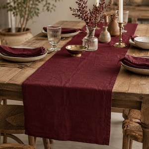 May include: A deep burgundy linen table runner laid across a rustic wooden dining table. The runner is paired with matching burgundy cloth napkins folded on ceramic plates, clear glassware, a small glass vase, and a brass candle holder.