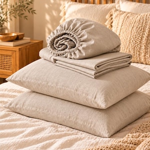 May include: A set of beige linen bedding including two rectangular pillows stacked on top of each other, a folded flat sheet, and a fitted sheet with an elasticized edge. The fabric has a visible woven texture in a neutral light tan color.