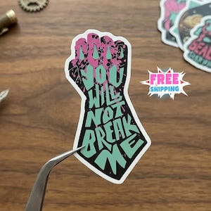 May include: A die-cut sticker shaped like a clenched fist, featuring a black background with pink splatter accents and the light green text "YOU WILL NOT BREAK ME" integrated into the design, held by a pair of silver metal tweezers.