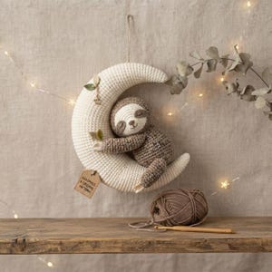 Peut inclure: Un croissant de lune crocheté couleur crème avec un paresseux marron accroché dessus. La lune comporte une petite breloque étoile, une étiquette marron avec le texte "HANDMADE A ORIGINAL PATTERN" et deux feuilles en tissu vert. Une pelote de laine et un crochet sont en dessous.