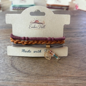 May include: A set of three stacked bracelets on a beige display card labeled Ember Hill Jewelry. The bracelets include a textured maroon cord, a braided mustard yellow cord, and a grey multi-strand cord featuring a gold leaf charm and bead.