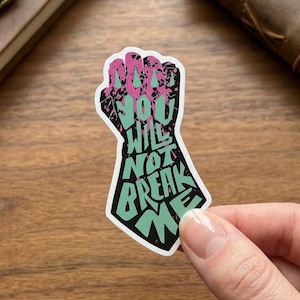 May include: A die-cut sticker shaped like a clenched fist, featuring a black background with a light green graphic of the text "YOU WILL NOT BREAK ME" and a pink, speckled, distressed texture overlaying the knuckles and upper portion.