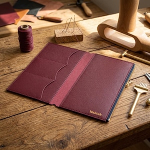 Könnte beinhalten: Ein bordeauxrotes Lederetui mit einem Reißverschlussfach und drei innenliegenden Kartenfächern. Auf der Außenseite befindet sich der gold geprägte Schriftzug Maktub. Das Etui ist geöffnet und zeigt eine strukturierte Oberfläche.