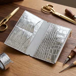 Op de afbeelding: Een zilveren portemonnee met een textuur van krokodillenleer, met een open interieur met kaartsleuven aan de linkerkant en een vak met ritssluiting aan de rechterkant, omringd door een gouden schaar en metalen gereedschappen.