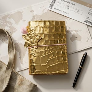 May include: A metallic gold notebook with a crocodile-embossed texture, secured by a light pink elastic band. It features a gold star-shaped charm and a small, fuzzy light pink pom-pom attached to the top corner of the cover.