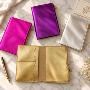 May include: Four rectangular passport holders in metallic finishes: one hot pink, one purple, one silver, and one open gold holder showing interior card slots. Each closed holder features a side zip with a small charm pull.