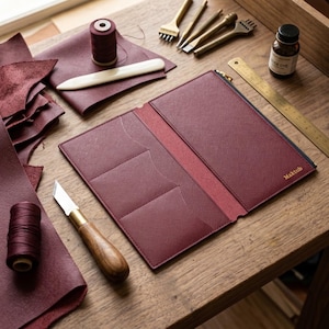 May include: A burgundy leather notebook cover with a zip closure, three interior card slots, and the word Maktub embossed in gold. The cover is surrounded by leather scraps, a spool of thread, a bone folder, a knife, and a metal ruler.