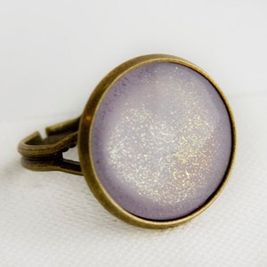 Pode incluir: Anel ajustável com uma grande face redonda de cor lavanda. A face do anel tem um design salpicado e brilhante com detalhes dourados e brancos. A banda e a moldura são de cor bronze.