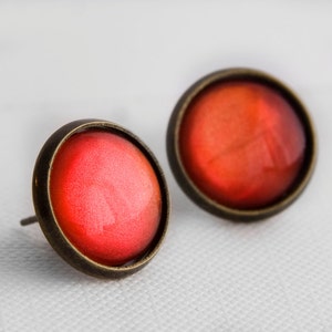 Könnte beinhalten: Ein Paar rote und bronzene Ohrstecker. Die Ohrringe sind rund, leicht gewölbt und haben eine glänzende, metallische Oberfläche.