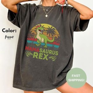 Pode incluir: Camiseta cor pimenta com um dinossauro verde com um laço vermelho, em um fundo de pôr do sol com palmeiras. O texto "Mamasaurus Rex" é impresso em verde e vermelho abaixo.