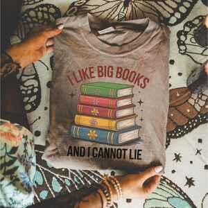 May include: A gray t-shirt with the text "I LIKE BIG BOOKS AND I CANNOT LIE" above a stack of colorful books. The books are green, red, pink, yellow, and blue, each with a flower design. The shirt is held by hands.