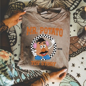 Può includere: Una maglietta color marrone chiaro con una grafica di Mr. Potato Head di Toy Story. Il design include il testo "MR. POTATO" in arancione, un cerchio a scacchi e le parole "TOY STORY" e "EST. 1999". La maglietta è piegata.