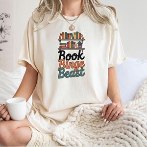 May include: Cream-colored t-shirt featuring a bookshelf graphic with colorful books and the text "Book Binge Beast" in black, orange, and teal. The shirt has short sleeves and a relaxed fit.