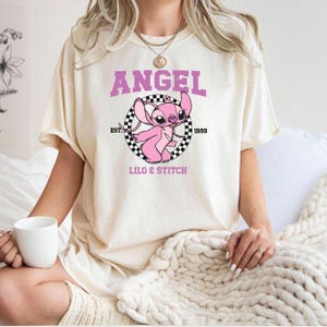 Pode incluir: Camiseta creme com um personagem de desenho animado rosa, Angel, de Lilo & Stitch. O design inclui a palavra "ANGEL" acima do personagem, com "EST. 1999" e "LILO & STITCH" abaixo. O personagem está em um círculo quadriculado em preto e branco.