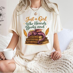 Pode incluir: Camiseta creme com o texto "Just a Girl Who Hoards Books and" acima de um dragão roxo descansando em uma pilha de livros. Ilustrações de penas douradas ladeiam o dragão.