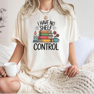 Pode incluir: Camiseta creme com o texto preto "I HAVE NO SHELF CONTROL". O gráfico inclui uma pilha de livros coloridos, uma vela acesa e um pequeno vaso de flores. Um estilo de camiseta casual e confortável.