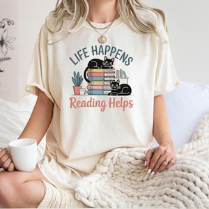 Puede incluir: Camiseta color crema con el texto "Life Happens Reading Helps" y un gráfico de dos gatos negros y una pila de libros. El diseño incluye una pequeña planta en maceta y una taza de café.