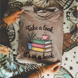 Puede incluir: Camiseta de color beige con la frase "Take a look, it's in a book" y un gráfico de libros, una taza de café y una planta. El gráfico es en negro, rosa, verde y amarillo. La camiseta está sujeta por una persona.