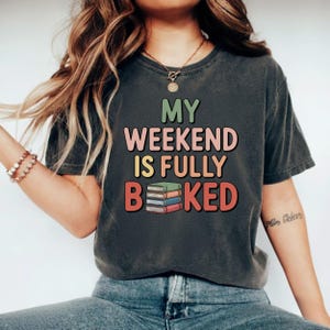 May include: Dark gray t-shirt with the text "MY WEEKEND IS FULLY BEKED" in colorful letters. The design includes a stack of books. A casual, comfortable tee for book lovers.