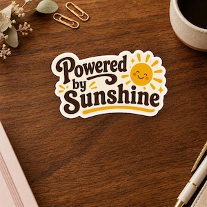 May include: A die-cut sticker featuring the dark brown text Powered by Sunshine in a bold, retro-inspired font. To the right of the text is a yellow cartoon sun with a smiling face, radiating small beams. The sticker has a white border.