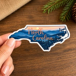 May include: A die-cut sticker shaped like the state of North Carolina featuring a scenic illustration of blue mountain ranges, a forest, and a sunset sky. The text North Carolina is printed in a cursive orange font across the center of the design.