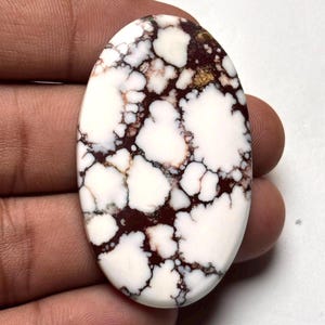 May include: An oval-shaped cabochon gemstone featuring a smooth, polished surface with a white base color and intricate, dark brown to black spiderweb matrix patterns throughout the stone, displaying a high-gloss finish and natural mineral veining.