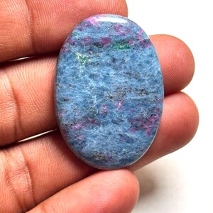 Op de afbeelding: Een ovaalvormige cabochon edelsteen met een textuurrijke blauwe basis, verspreid met insluitingen van levendig magenta en kleine groene vlekjes. Het oppervlak van de steen is gepolijst en glad, met een natuurlijk mineraalpatroon.