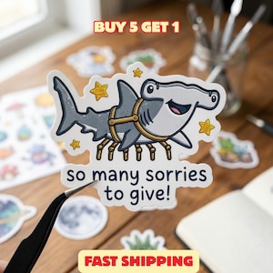 May include: A die-cut sticker featuring a cartoon hammerhead shark with spider-like legs, wearing a harness, surrounded by yellow stars. The text reads: BUY 5 GET 1, so many sorries to give!, and FAST SHIPPING. The sticker has a white border.