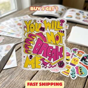 May include: A die-cut sticker featuring the text YOU WILL NOT BREAK ME in bold yellow and pink lettering. The design includes small illustrations of a dinosaur, a skull, a trophy, and a cat face, all set against a white background.
