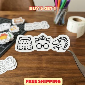 May include: A white die-cut sticker featuring three black pixel art icons arranged horizontally: a pair of shorts patterned with small hearts, a tiara resting above round glasses, and the profile of a dinosaur head with a spiky crest.