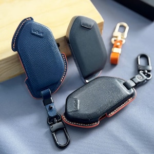 May include: Two textured leather key fob covers in blue and black, each featuring orange stitching and a black metal clasp. A plain black key fob and a small orange leather keychain with a gold-tone metal clip are also included in the display.