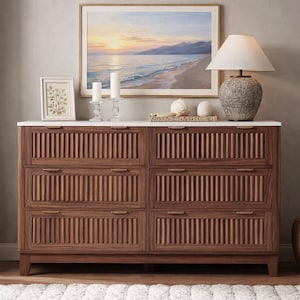 May include: A wooden dresser with six drawers, featuring vertical grooved detailing on the drawer fronts. The dresser has a white marble top and brass handles. A framed landscape painting, a lamp, and decorative items sit on top.
