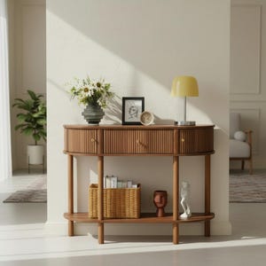 May include: A wooden console table with a ribbed front design, two drawers, and a lower shelf. The table is adorned with a vase of flowers, a framed picture, a small clock, a yellow lamp, a woven basket, and decorative sculptures. The table is made of wood.