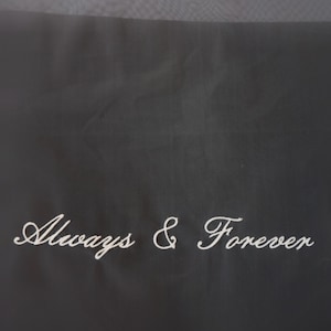 May include: A black fabric surface featuring the white embroidered text Always & Forever in a cursive script font. The material has a smooth, matte texture with a subtle weave visible across the dark background, highlighting the white lettering.