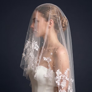 May include: A sheer white bridal veil featuring delicate floral lace appliqués along the edges and scattered small white pearls throughout the tulle fabric. The veil drapes over the head and shoulders, showcasing intricate embroidery and beadwork.