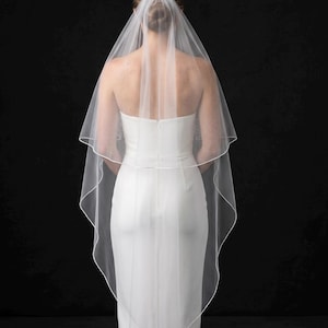 May include: A two-tier white bridal veil featuring a delicate pencil edge trim. The sheer tulle fabric is layered, with the shorter top tier cascading over the longer bottom tier, creating a soft, flowing silhouette against a solid black background.