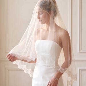 May include: A sheer, ivory-colored bridal veil featuring delicate, scalloped lace trim along the edges. The veil is crafted from fine, lightweight tulle fabric that drapes softly over the shoulders, creating a classic and elegant appearance.