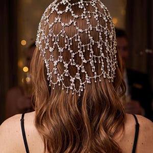 May include: A silver-toned headpiece featuring a delicate, net-like chain structure adorned with numerous small, clear, sparkling crystals. The accessory drapes over the top and back of the head, creating a cascading, layered effect with dangling crystal accents.