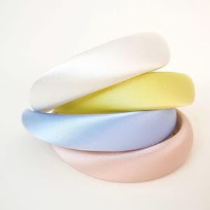 May include: A stack of four padded headbands in pastel colors. The top headband is white, followed by a yellow one, a light blue one, and a pale pink one at the bottom. Each headband has a smooth, satin-like finish and a rounded, thick profile.