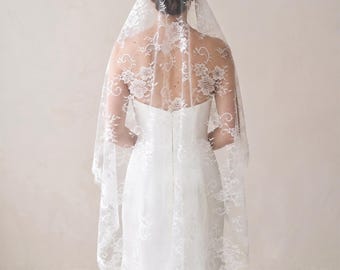 May include: A white lace veil featuring an intricate floral pattern with scalloped edges, draped over the head and shoulders. The sheer fabric displays detailed embroidery of blossoms and swirling vines, extending down the back in a soft, flowing shape.