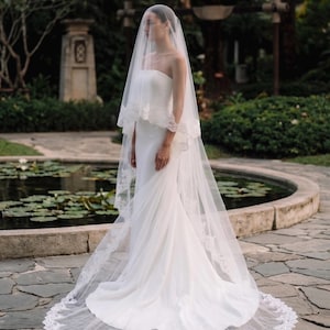 May include: A floor-length white strapless wedding dress with a smooth, fitted bodice and a flowing skirt. The ensemble includes a long, sheer tulle veil that drapes over the head and shoulders, featuring intricate white lace trim along the edges.